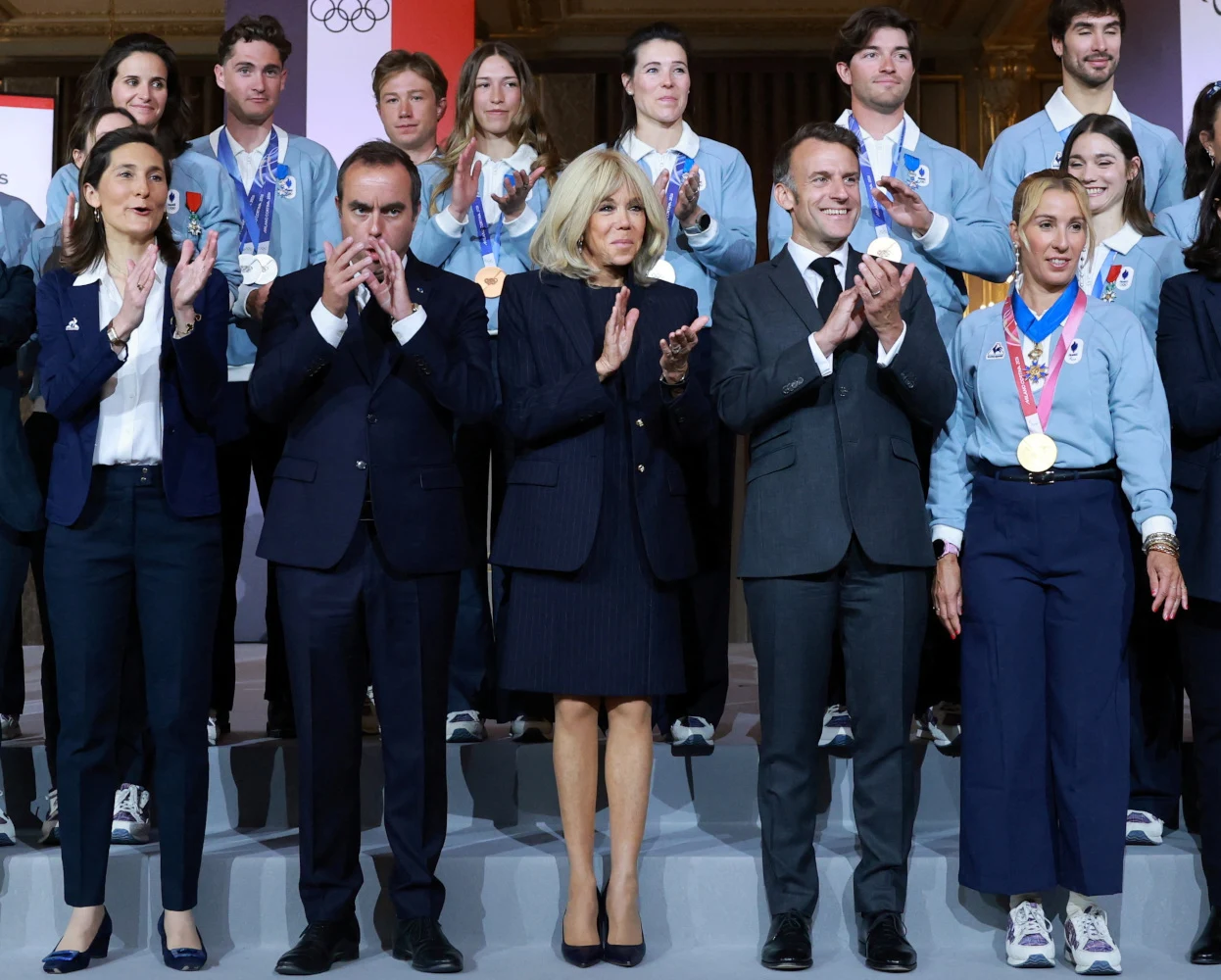 Grupa sportowców ubranych w oficjalne stroje stoi na podium, w pierwszym rzędzie wyróżniają się cztery osoby w eleganckich garniturach oraz jedna kobieta z trzema medalami na szyi, wszyscy klaszczą i patrzą w stronę publiczności.