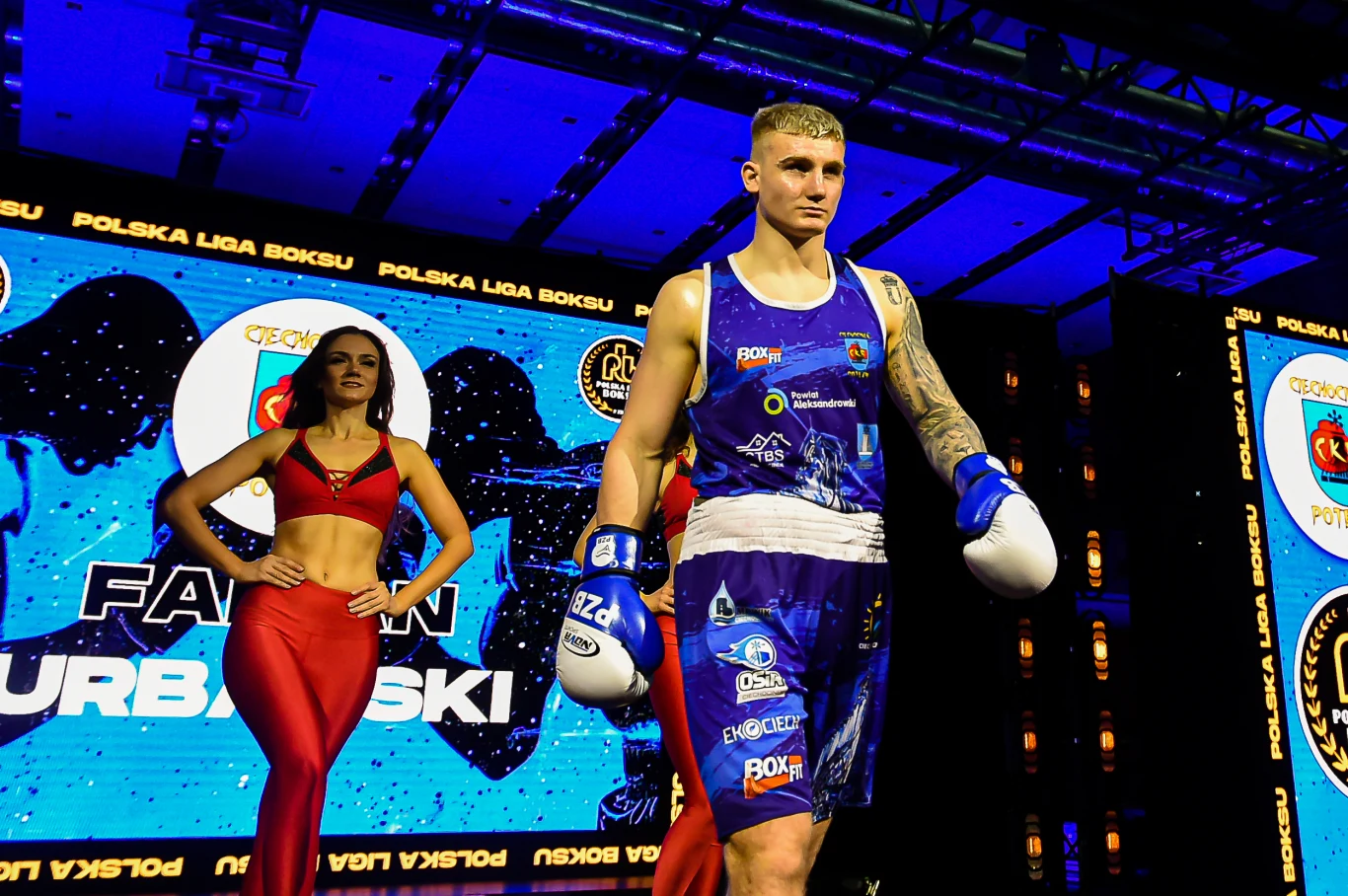 Fabian Urbański ma wszystko ku temu, aby zostać znakomitym pięściarzem w gronie seniorów bokser ubrany w niebiesko-białe spodenki i rękawice wychodzi na ring, obok niego stoi hostesska w czerwonym stroju, w tle rozświetlona scena z dużym ekranem prezentującym nazwiska i logotypy związane z boksem