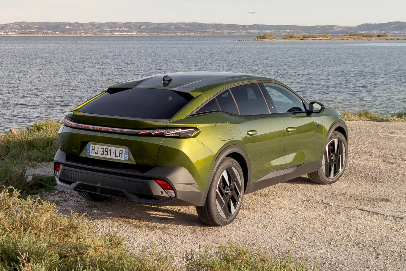 Emblemat z lwem został zastąpiony podświetlanym napisem "Peugeot". zielony samochód typu crossover zaparkowany na żwirowym terenie nad brzegiem jeziora, w tle panorama niskich wzgórz i wody