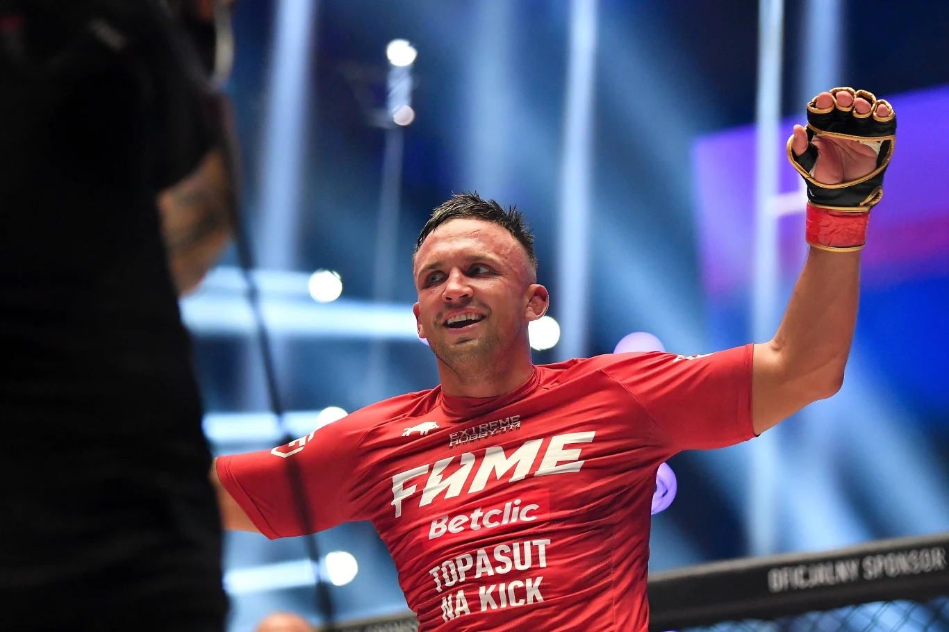 Robert Pasut zawodnik w czerwonej koszulce triumfalnie unosi ręce w górze w oktagonie, na twarzy ma szeroki uśmiech, w tle widać światła areny i elementy klatki