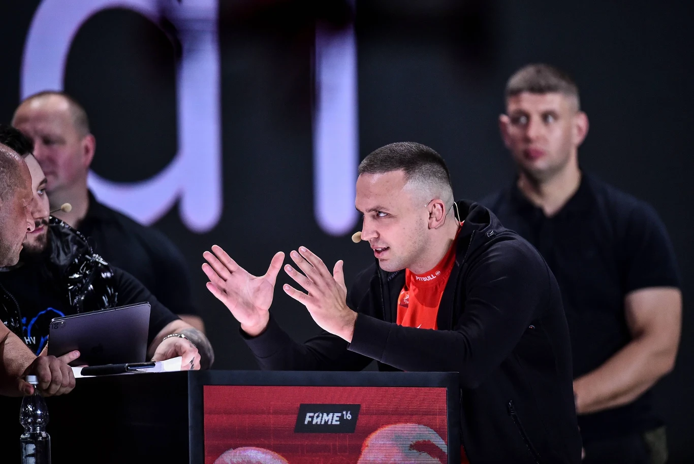 Robert Pasut Mężczyzna w czarnej bluzie intensywnie gestykuluje, stojąc za pulpitem z napisem 'FAME 16', obok dwóch innych mężczyzn, z których jeden trzyma notes, a drugi stoi z rękami skrzyżowanymi na tle ciemnego, nieostrego napisu.