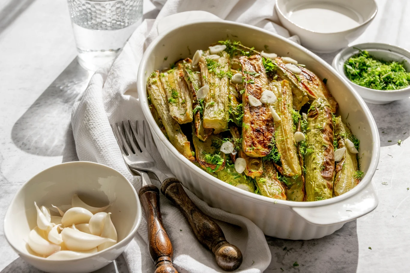 Grillowana młoda cukinia lub szparagi to świetny dodatek do grillowanych warzyw. Pełen błonnika Zapiekane plastry cukinii ułożone w białym naczyniu żaroodpornym, posypane świeżymi ziołami i plasterkami czosnku, w otoczeniu miseczek z dodatkami i szklanki wody.
