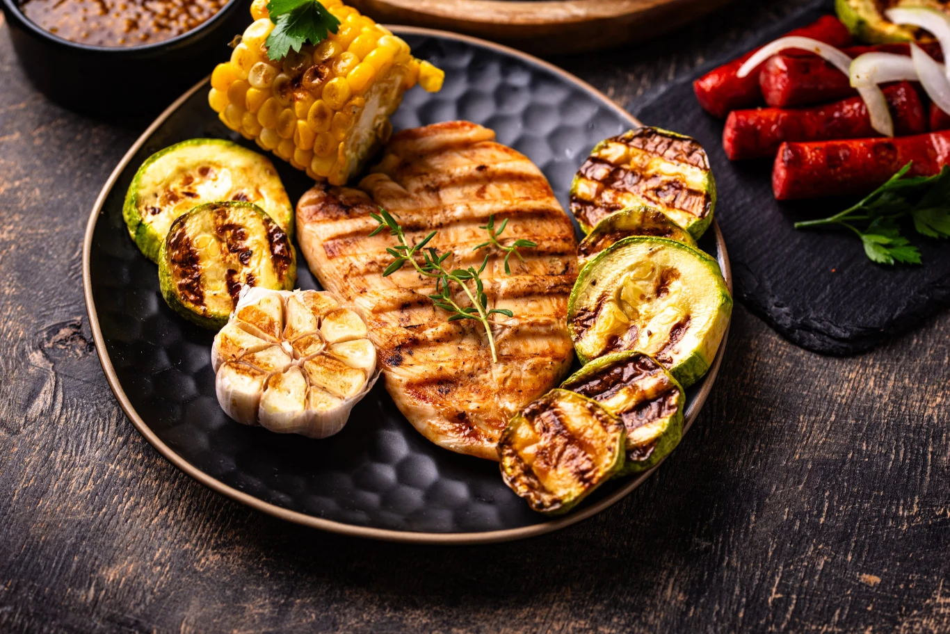 Lubisz grillowane mięso? Ok, ale nie pomijaj źródeł błonnika Grillowana pierś z kurczaka podana z grillowaną cukinią, kukurydzą i opiekaną główką czosnku, udekorowana świeżym tymiankiem, na czarnym talerzu.