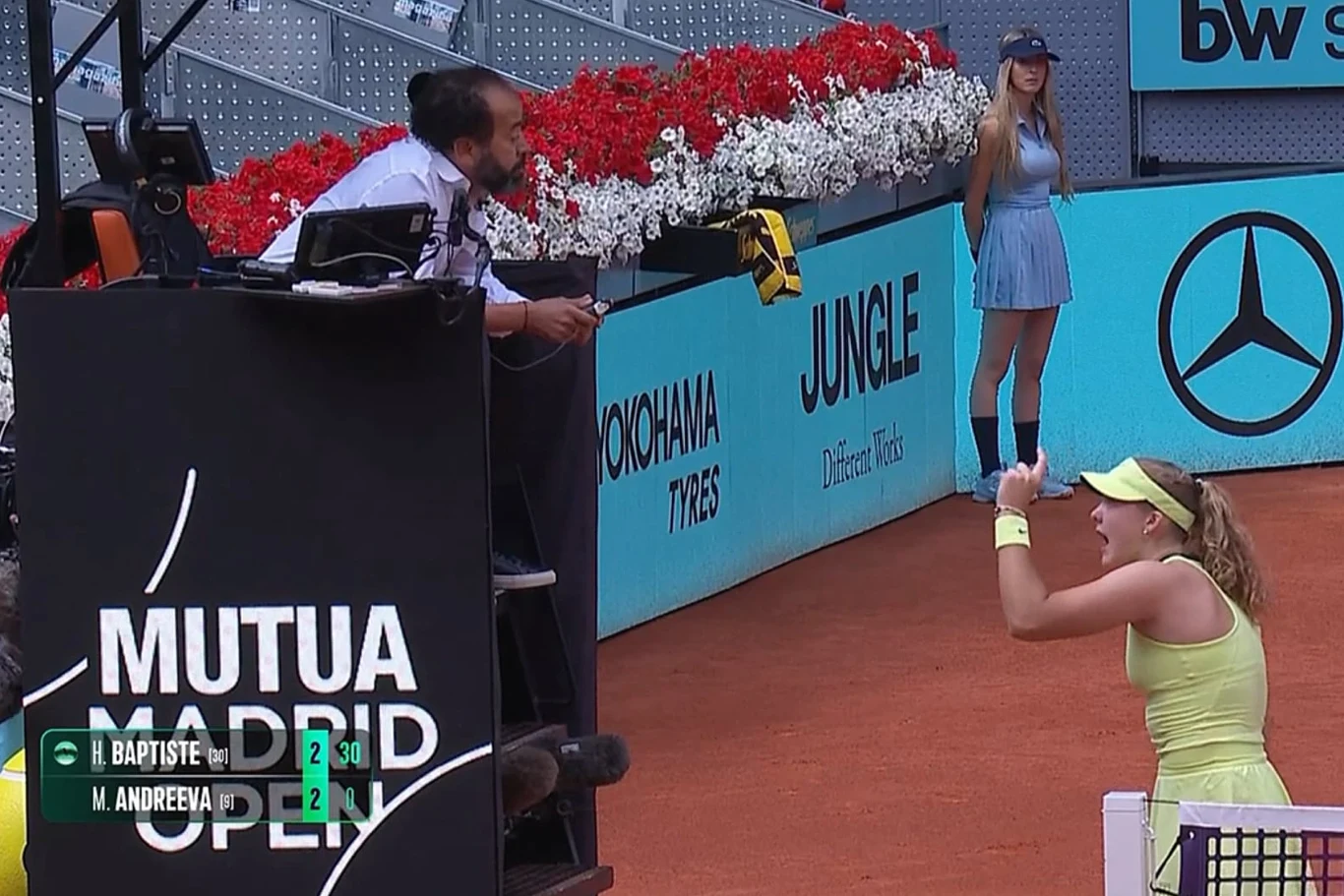 tenisistka w jasnożółtym stroju gestykuluje i rozmawia z sędzią siedzącym na fotelu przy korcie, na tle czerwonych kwiatów w alejce reklamowej, z tyłu stoi ball girl w błękitnej sukience, widoczny wynik meczu to 2:2 w gemach
