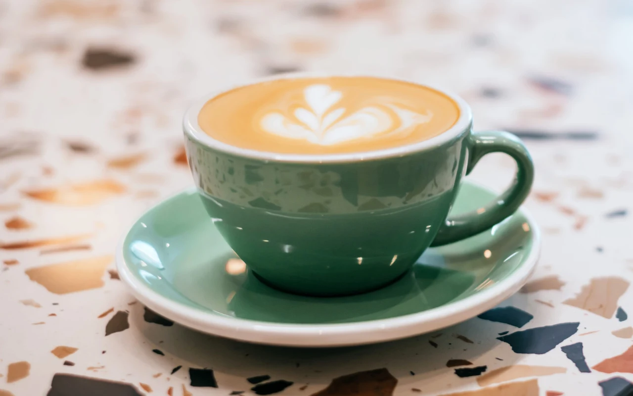 zielona filiżanka z latte na spodku, na powierzchni kawy wzór wykonany mleczną pianką, ustawiona na stoliku z dekoracyjnym, mozaikowym blatem