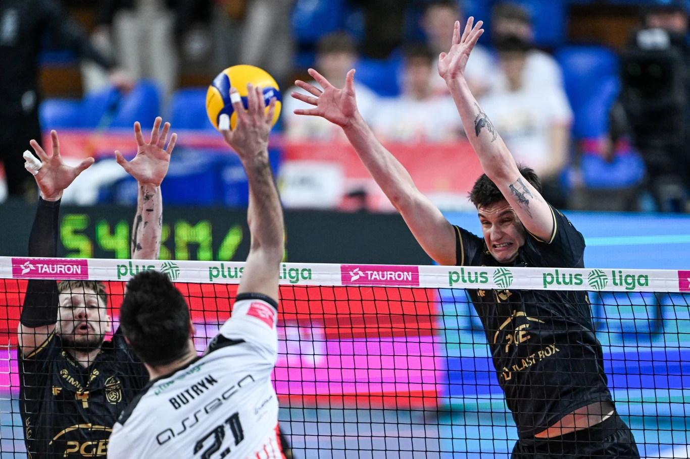 dwóch siatkarzy drużyny w czarnych strojach próbuje zablokować atak zawodnika w białym stroju, piłka znajduje się tuż nad siatką, atmosfera rywalizacji sportowej podkreślona logotypami ligowymi na bandach
