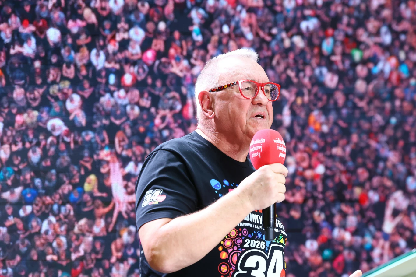 Jurek Owsiak razem z pozostałymi jurorami wybrał 16 finalistów Eliminacji do Pol'and'Rock Festivalu Jurek Owsiak z mikrofonem, w okularach i czarnej koszulce na tle tłumu podczas Eliminacji Pol'and'Rock Festivalu