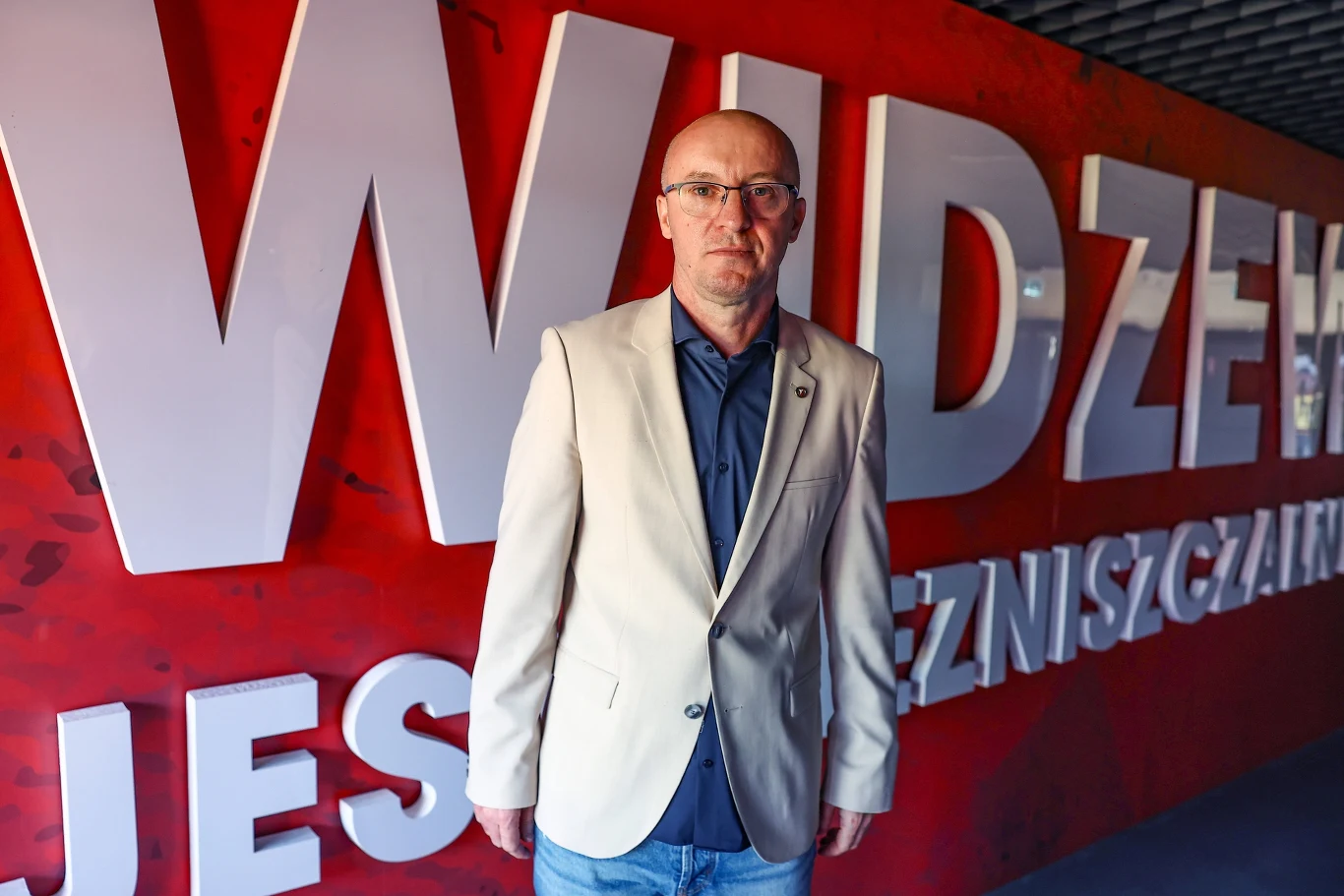 Aleksandar Vuković od marca jest trenerem Widzewa Aleksandar Vuković w okularach, jasnej marynarce i ciemnej koszuli na tle czerwonej ściany z napisem 'WIDZEW'.