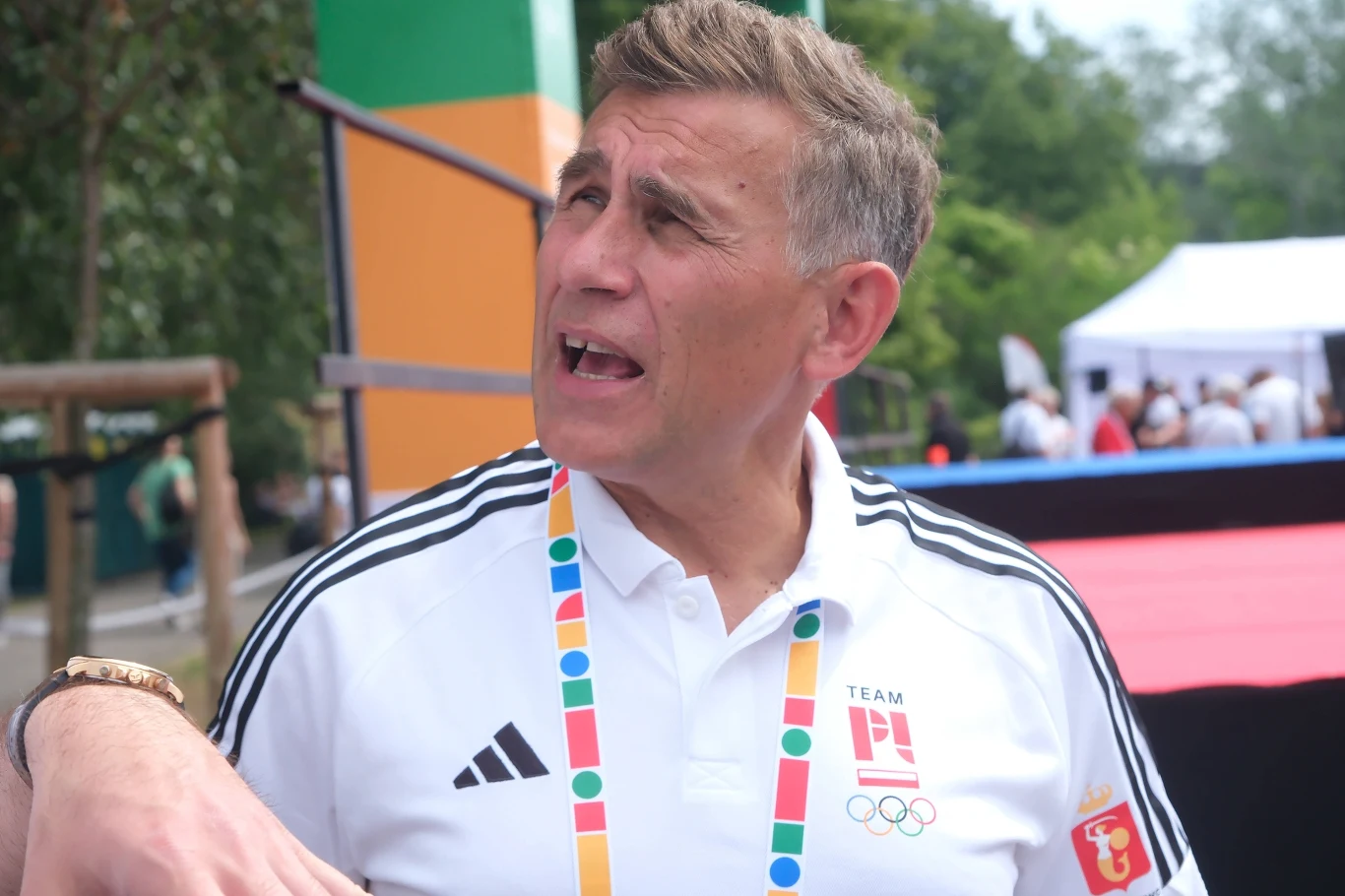 mężczyzna w białej koszulce z logo drużyny olimpijskiej i kolorową smyczą na szyi, stoi na tle obiektów sportowych na świeżym powietrzu
