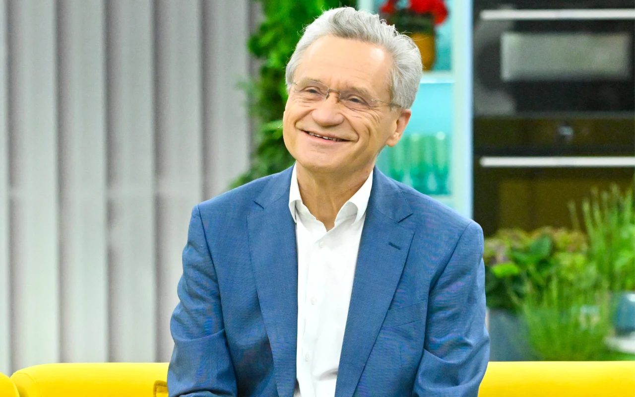 Mężczyzna w niebieskiej marynarce i białej koszuli siedzi na żółtej sofie, uśmiecha się szeroko, w tle widoczna dekoracja z roślinami oraz półki z przedmiotami.