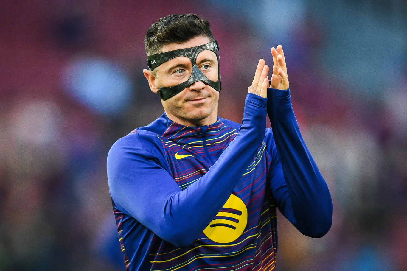 Piłkarz w sportowej koszulce z herbem i logo sponsora ma na twarzy czarną maskę ochronną i składa dłonie do oklasków, stojąc na tle rozmytego stadionu.