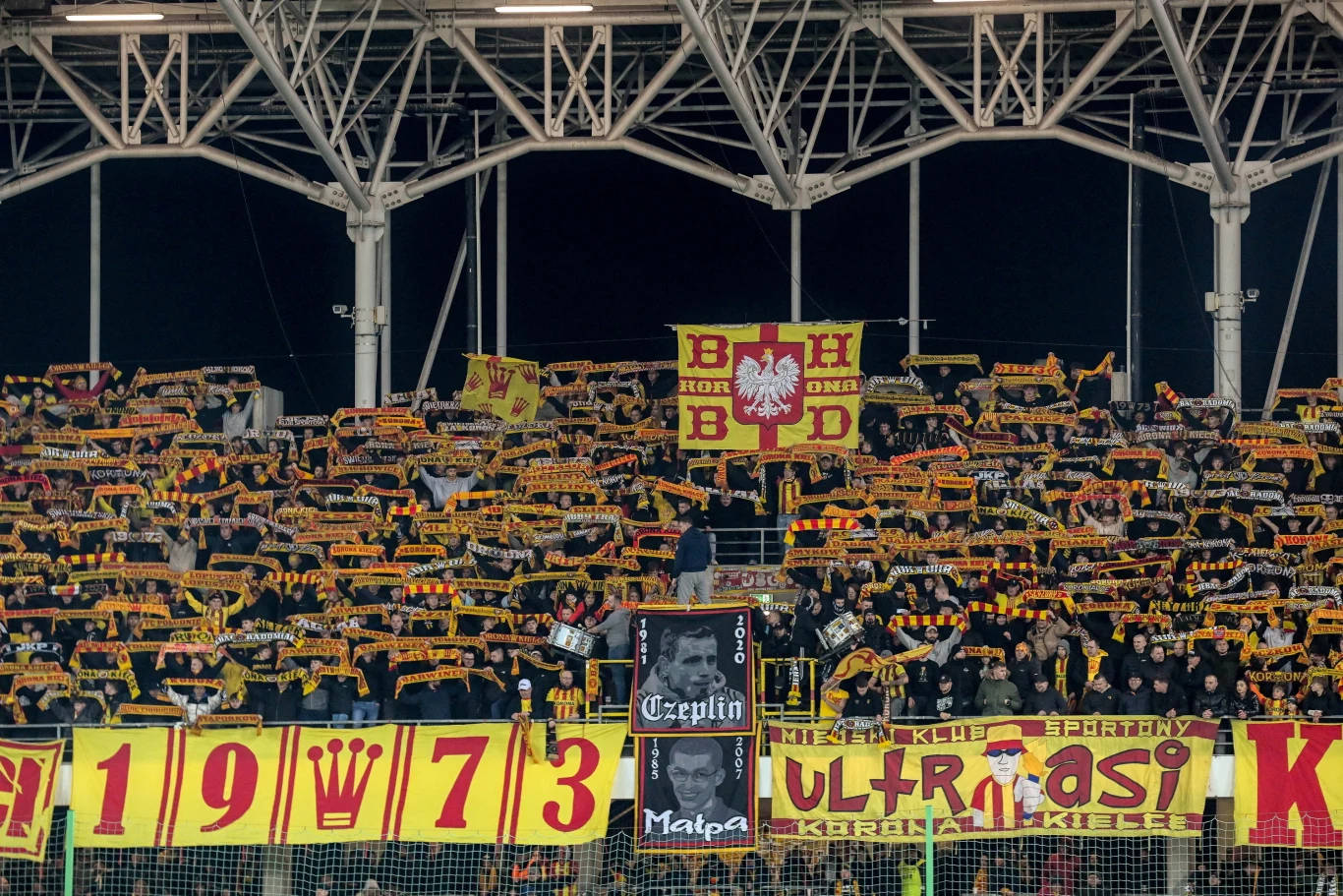 Kibice Korony Kielce tłum kibiców na trybunach stadionu unosi żółto-czerwone szaliki, nad głowami widoczne liczne transparenty i flagi z napisami oraz symbolem biało-czerwonej Polski, na dole duży baner z datą 1973 i napisem 'ultrasi'
