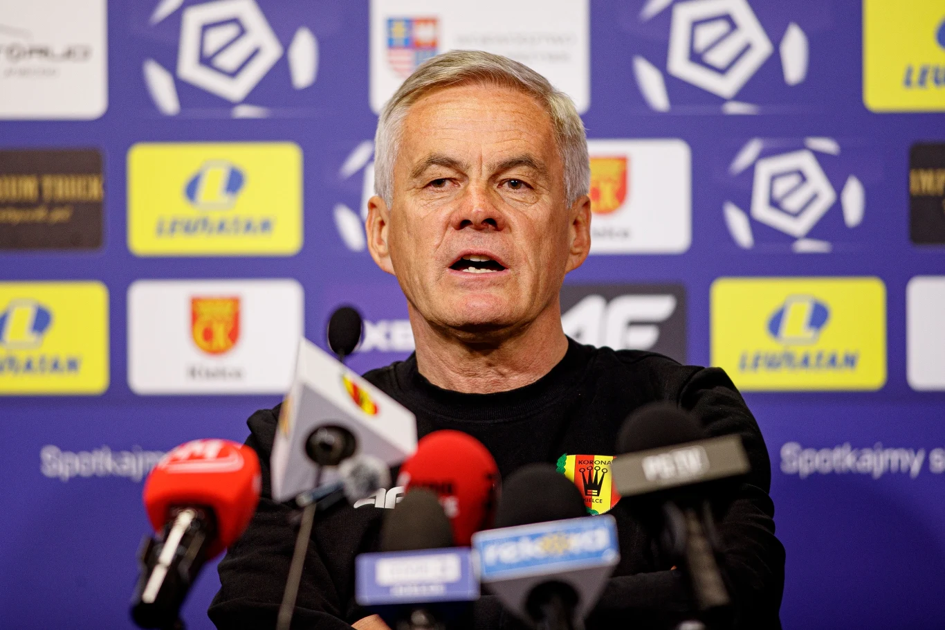 Jacek Zieliński, trener Korony Kielce Mężczyzna o siwych włosach udziela wywiadu na konferencji prasowej, siedząc przed stołem z kilkoma mikrofonami różnych stacji, na tle ścianki z logotypami sponsorów i ligi piłkarskiej.