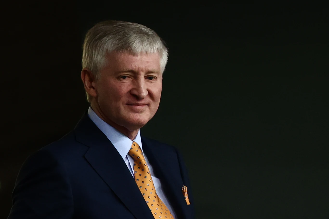 Rinat Achmetow starszy mężczyzna w eleganckim granatowym garniturze z białą koszulą i żółtym krawatem w delikatne wzory, patrzący lekko w bok na tle ciemnego, jednolitego tła
