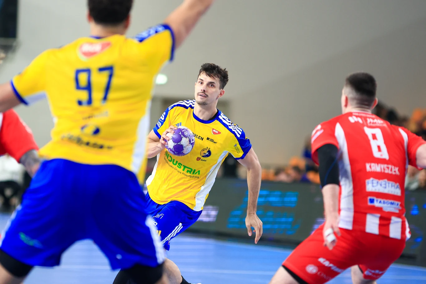 Industria Kielce po raz kolejny w sezonie zagra z Rebud KPR Ostrovią Ostrów Wielkopolski zawodnik w żółto-niebieskim stroju z piłką ręczną w dłoni atakuje bramkę, otoczony przez dwóch rywali w czerwono-białych strojach, w dynamicznej akcji podczas meczu piłki ręcznej