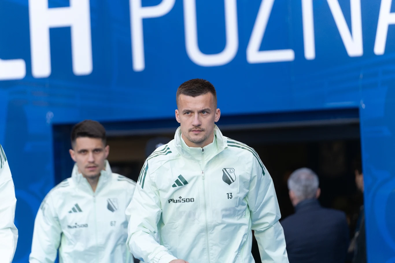 Piłkarze Legii Warszawa w jasnych dresach wychodzą tunelu stadionu przed meczem, na tle niebieskiego napisu 'Poznań'.