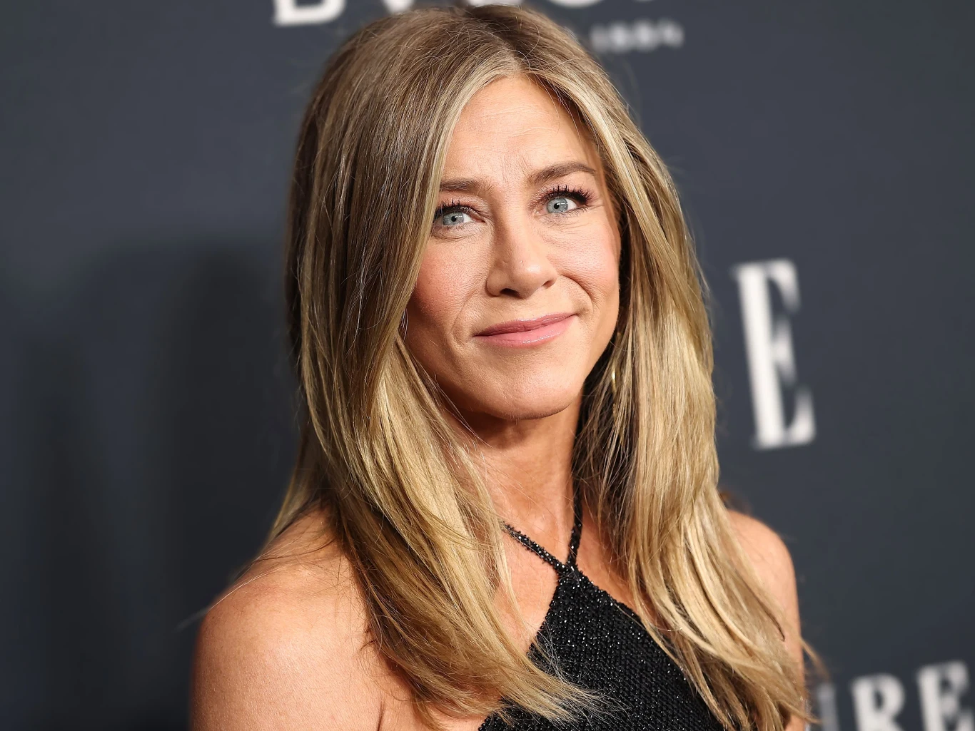 Jennifer Aniston Kobieta z długimi, jasnobrązowymi włosami ubrana w elegancką, czarną suknię stojąca na tle ścianki z dużymi białymi literami