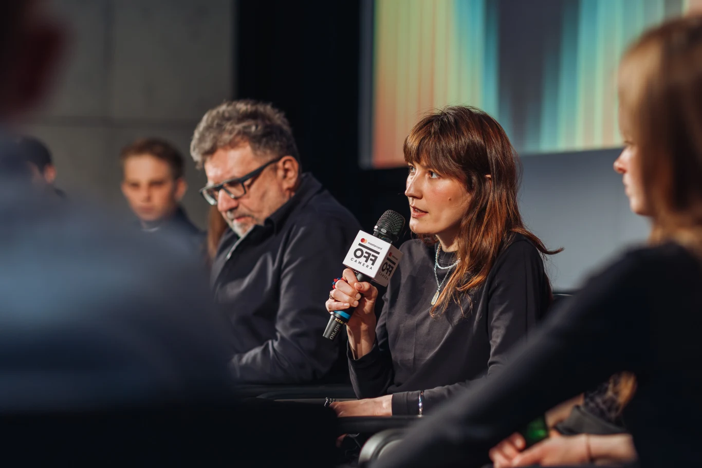 Emi Buchwald Kobieta trzymająca mikrofon z logo siedzi wśród kilku osób na tle kolorowego ekranu podczas panelu dyskusyjnego