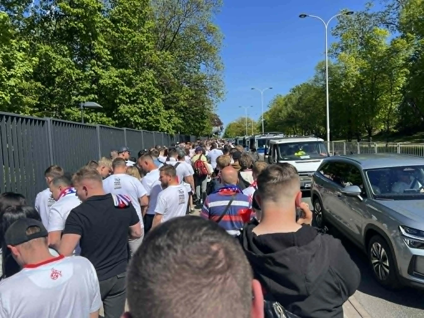 Tłumy przed PGE Narodowym duża grupa ludzi ubranych w białe koszulki i szaliki sportowe stoi w kolejce na chodniku obok ulicy, po prawej stronie widoczne są zaparkowane samochody, a w tle drzewa i jasne niebo
