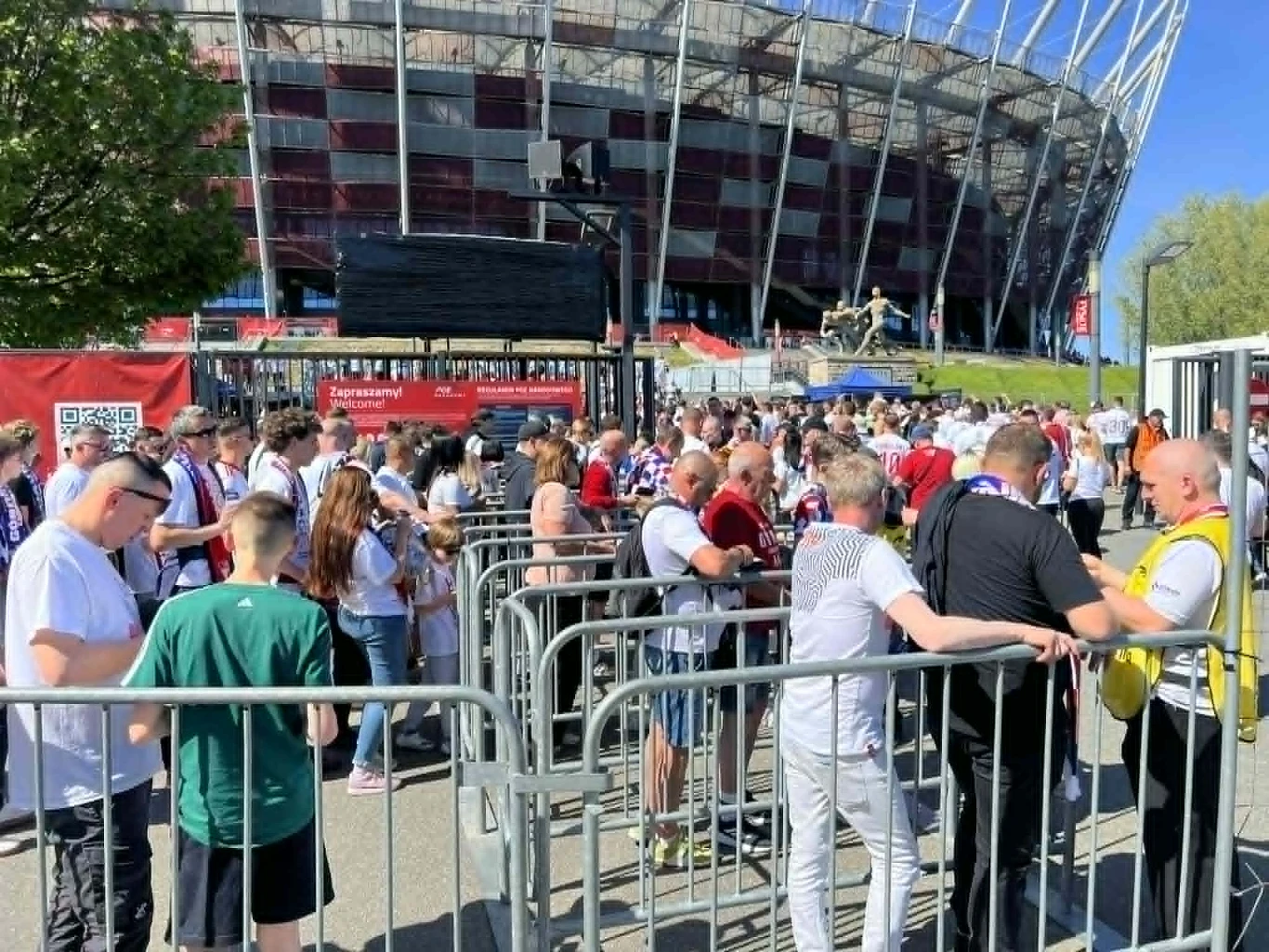 Tłumy przed PGE Narodowym Tłum ludzi ustawionych w kolejce przed wejściem na stadion, większość ubrana w biało-czerwone stroje kibicowskie, kontrola biletów przez pracownika ochrony, w tle widoczna bryła stadionu z charakterystyczną elewacją i bezchmurne niebo.