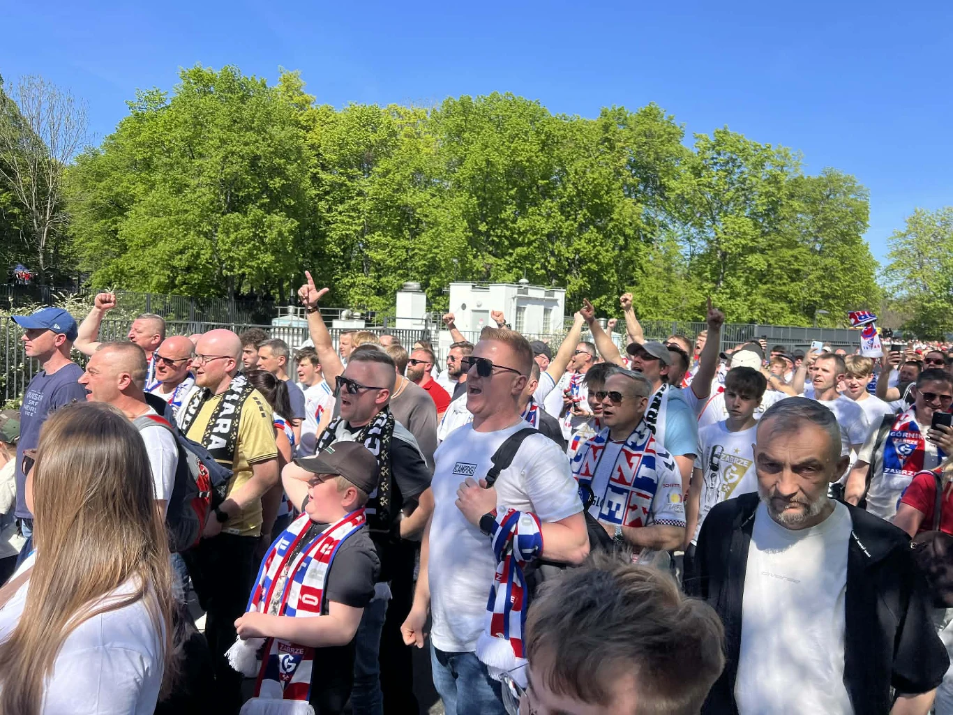 Tłumy przed PGE Narodowym tłum kibiców ubranych w barwy klubowe, z szalikami i koszulkami, idących razem w stronę stadionu, wielu z podniesionymi rękami i wyrazem entuzjazmu na twarzach, w tle zielone drzewa i jasne niebo