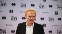 mężczyzna w średnim wieku w eleganckim garniturze i białej koszuli stoi na tle ścianki z logo festiwalu OFF Camera oraz innymi znakami graficznymi, patrząc lekko w bok