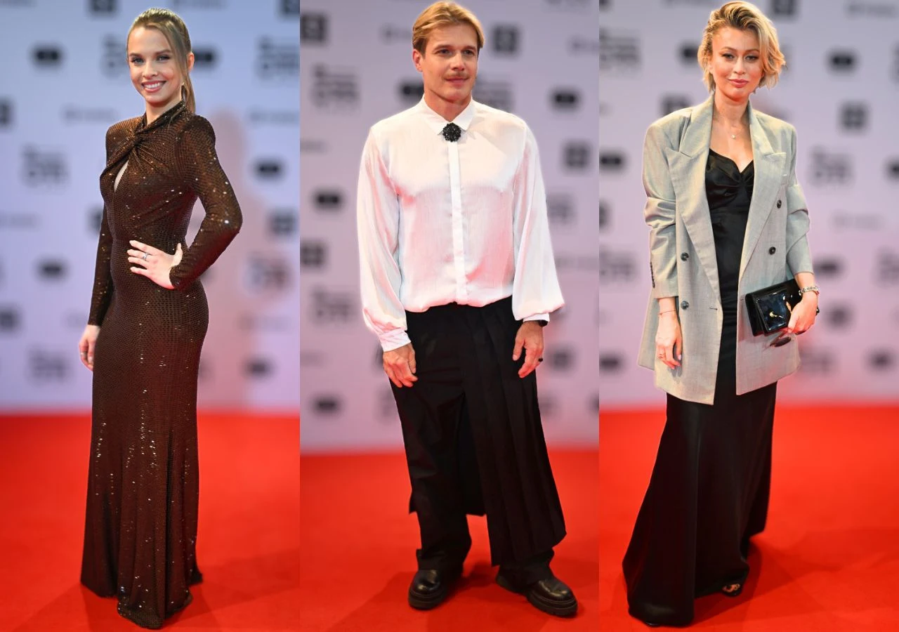 trzy osoby pozują na czerwonym dywanie w eleganckich stylizacjach wieczorowych; pierwsza z lewej ma na sobie długą błyszczącą suknię z długim rękawem, środkowa osoba ubrana jest w białą koszulę z szerokimi rękawami oraz czarną, luźną spódnicę i buty, n...