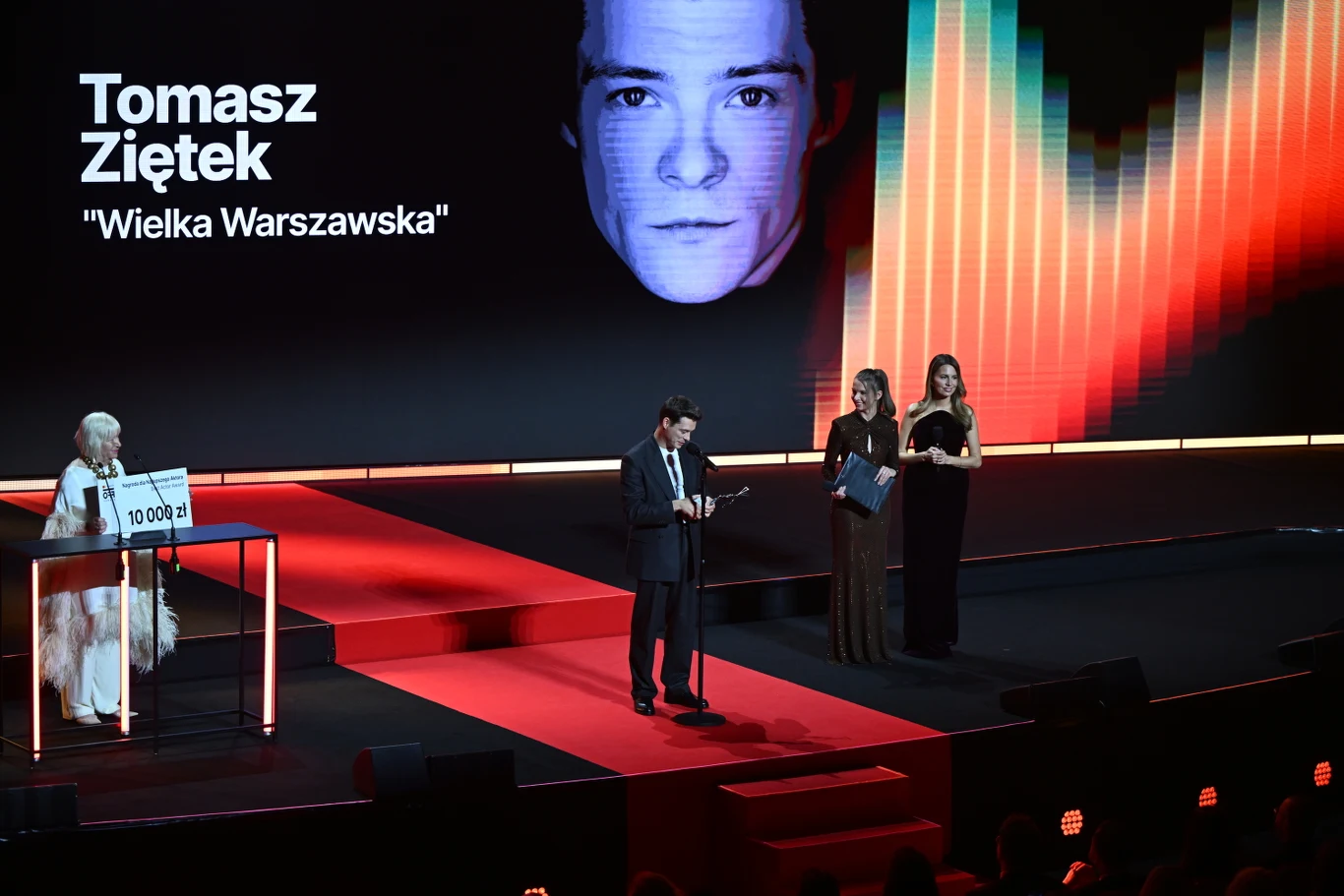 Tomasz Ziętek z nagrodą dla najlepszego aktora 19. edycji festiwalu Mastercard OFF CAMERA scena gali wręczenia nagród z dużym ekranem w tle wyświetlającym nazwisko Tomasza Ziętka oraz tytuł „Wielka Warszawska”; mężczyzna w garniturze stoi przy mikrofonie na środku sceny, po lewej kobieta trzyma tabliczkę z napisem „10 000 zł”, obok niej ora...