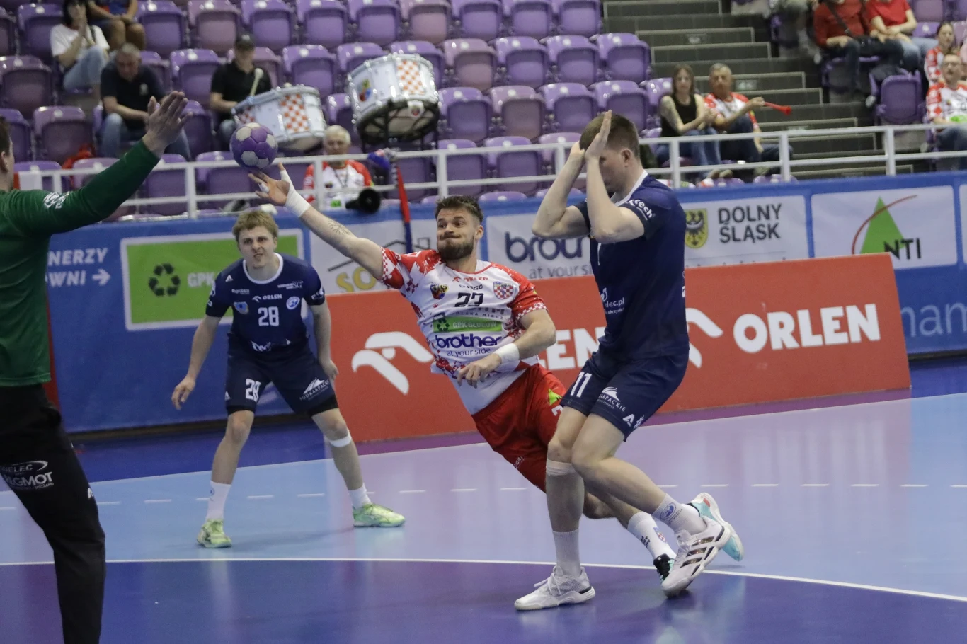 KGHM Chrobry Głogów – Handball Stal Mielec dwóch zawodników rywalizuje o piłkę ręczną w dynamicznej akcji pod bramką, jeden w biało-czerwonym stroju rzuca piłkę w kierunku bramki, blokowany przez przeciwnika w niebieskim stroju; w tle widoczni inni zawodnicy, trybuny oraz reklamowe banery