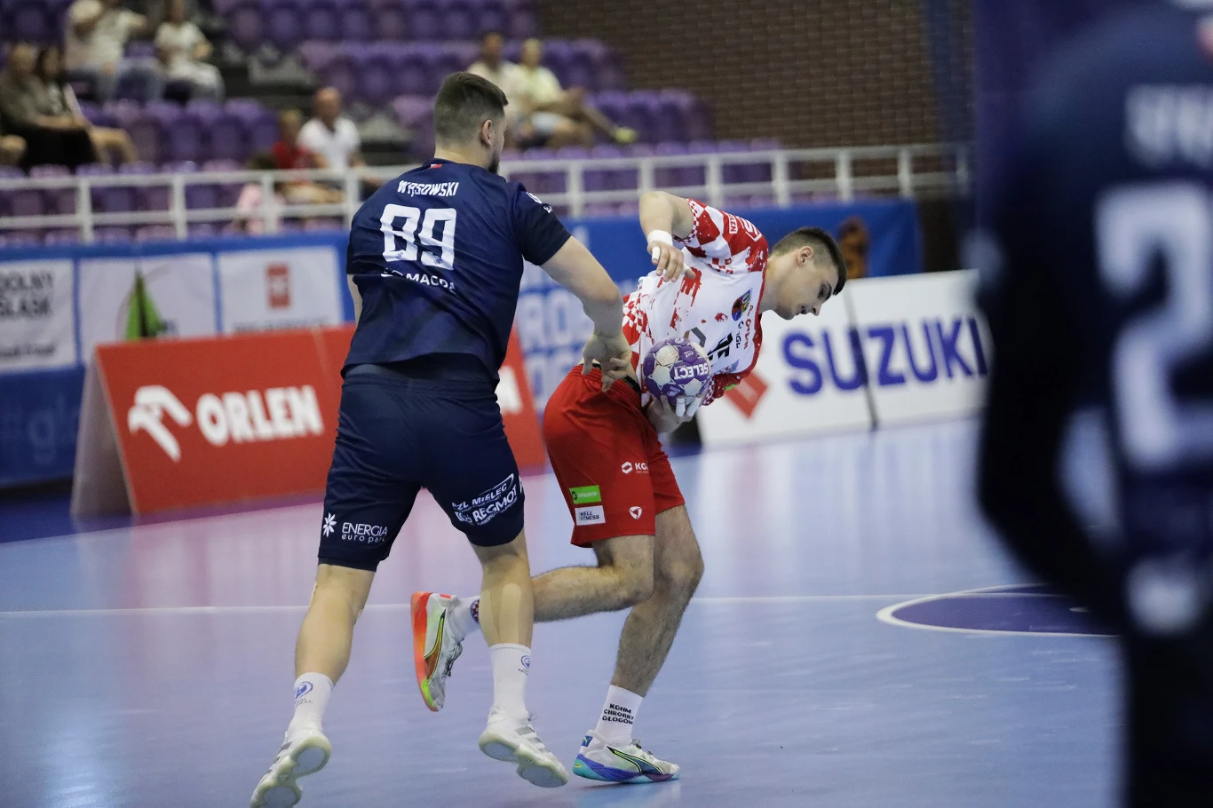 KGHM Chrobry Głogów – Handball Stal Mielec zawodnik w granatowym stroju z numerem 89 zatrzymuje zawodnika w biało-czerwonym stroju podczas dynamicznej akcji meczu piłki ręcznej na hali sportowej