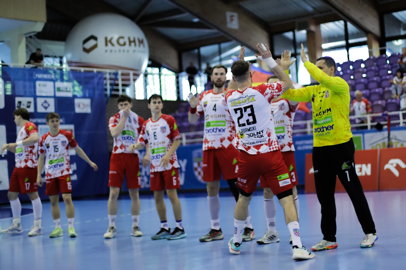KGHM Chrobry Głogów – Handball Stal Mielec zawodnicy drużyny piłki ręcznej ubrani w biało-czerwone stroje wykonują gesty powitania na boisku, w tle widoczne są trybuny z fioletowymi krzesełkami oraz banery sponsorów