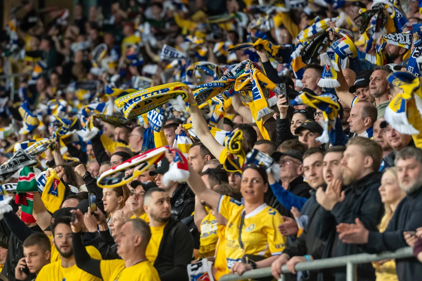 Tłum kibiców ubranych w żółte koszulki i szaliki klubowe unosi ręce, wiwatuje i okazuje wsparcie swojej drużynie podczas meczu wypełniając trybuny stadionu żywiołową energią.