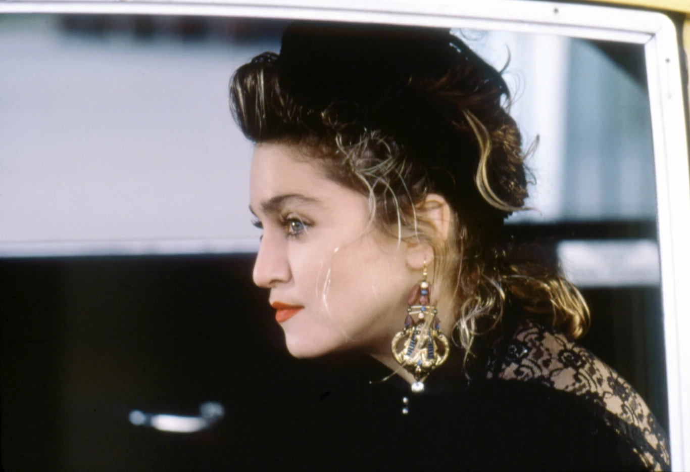 Madonna w filmie "Rozpaczliwie poszukując Susan" Madonna jako Susan w filmie, z wyrazistym makijażem, dużymi kolczykami i kręconymi włosami, patrzy przez okno auta