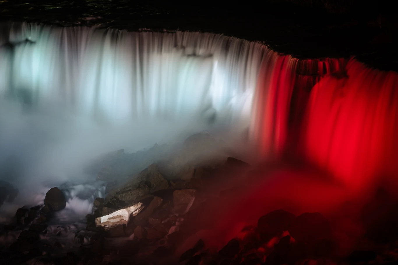 Wodospad Niagara podświetlono na biało-czerwone barwy Wodospad oświetlony intensywnym światłem w kolorach białym i czerwonym spływa kaskadami na ciemne, skaliste podłoże