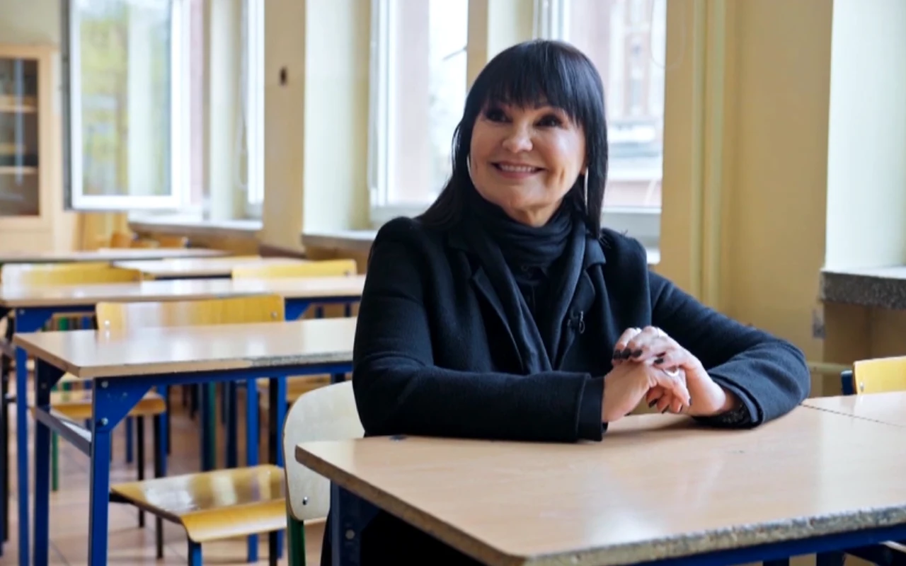 Kobieta z ciemnymi włosami i grzywką, ubrana na czarno, siedzi uśmiechnięta przy szkolnej ławce w pustej sali lekcyjnej.