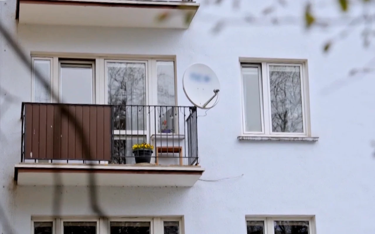 Balkon w bloku mieszkalnym z metalową balustradą, kwiatkiem w doniczce oraz zamontowaną talerzem anteny satelitarnej na jasnej elewacji budynku.