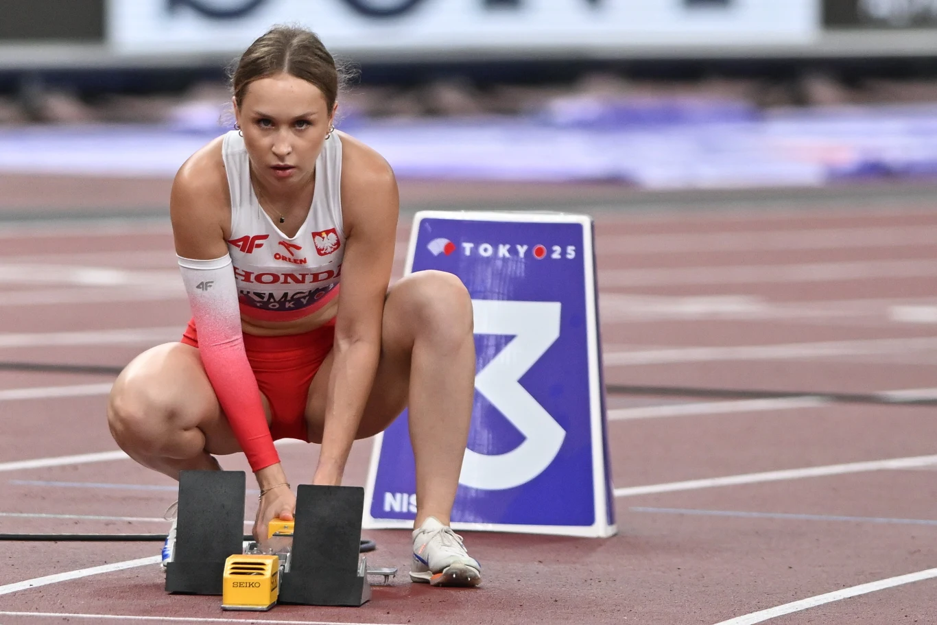 zawodniczka w stroju reprezentacji Polski przygotowująca się do startu biegu na stadionie lekkoatletycznym, klęcząca przy blokach startowych na torze numer 3, skupiona przed rozpoczęciem wyścigu