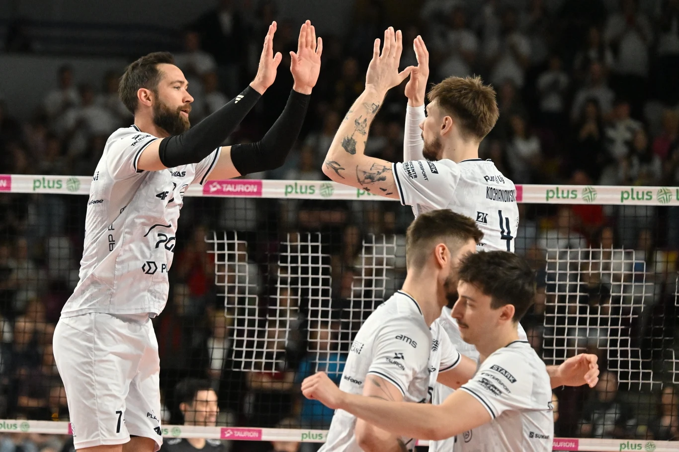 Siatkarze w białych strojach świętują udaną akcję przy siatce, przybijając sobie piątki na tle tłumu kibiców na hali sportowej.