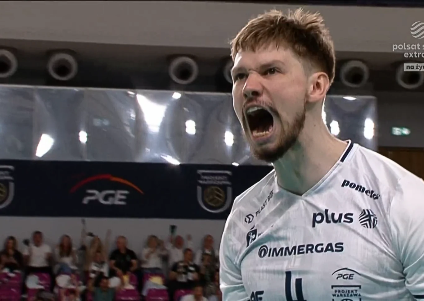 siatkarz w białym stroju z numerem 4 wykrzykuje z wielkimi emocjami, w tle rozmyte sylwetki kibiców oraz elementy hali sportowej