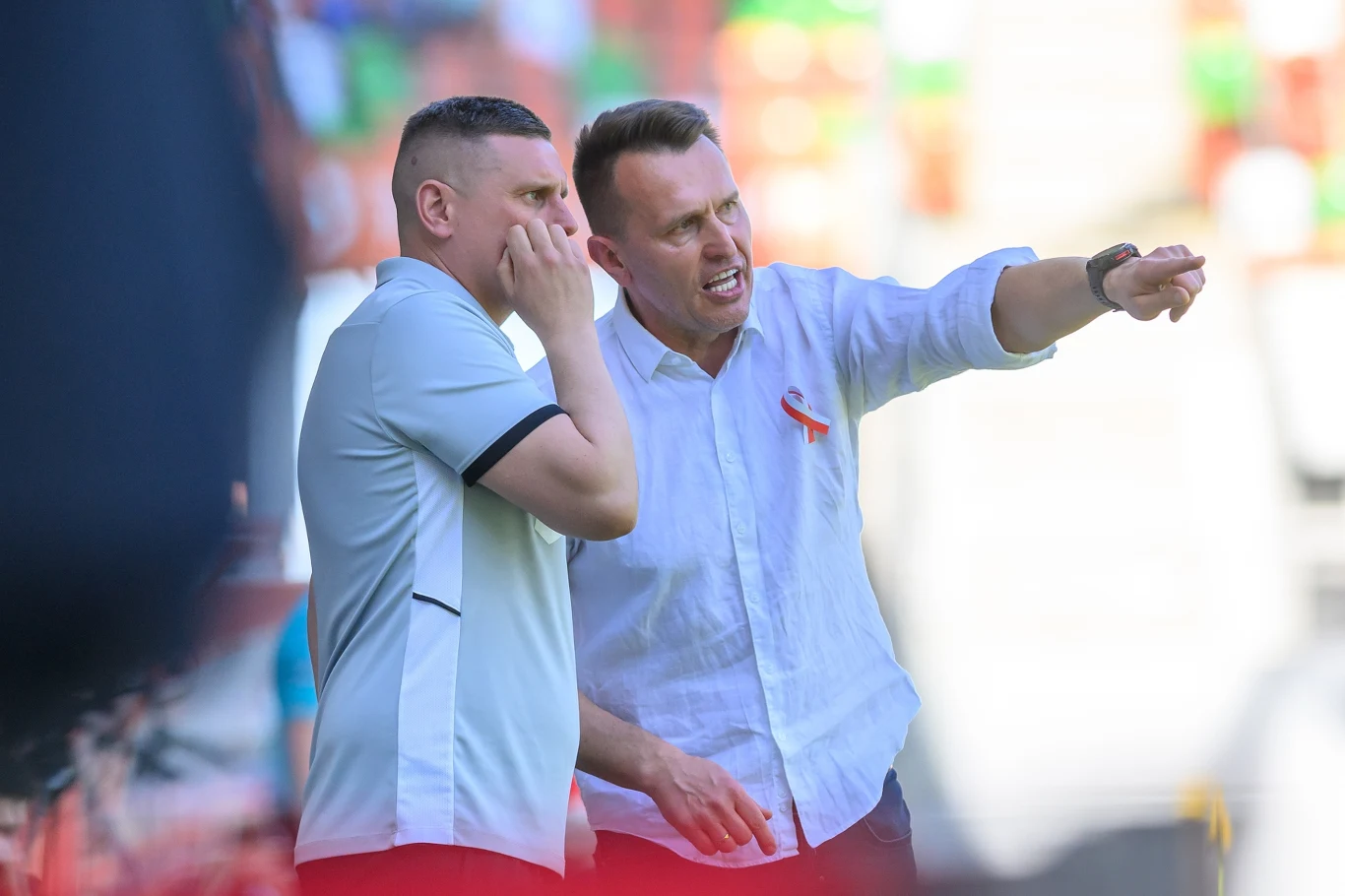 Dwóch mężczyzn w sportowych ubraniach dyskutuje, jeden z nich wskazuje ręką kierunek, obaj sprawiają wrażenie zaangażowanych w rozmowę na boisku sportowym.