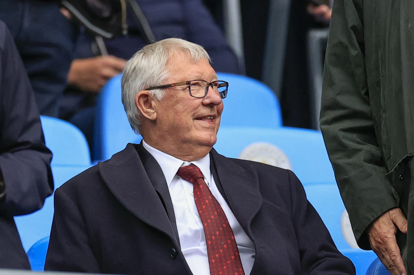 Starszy mężczyzna w okularach, ubrany w elegancki płaszcz i czerwoną krawatę, siedzi na niebieskim stadionowym krześle, uśmiechając się i rozmawiając z osobą obok.
