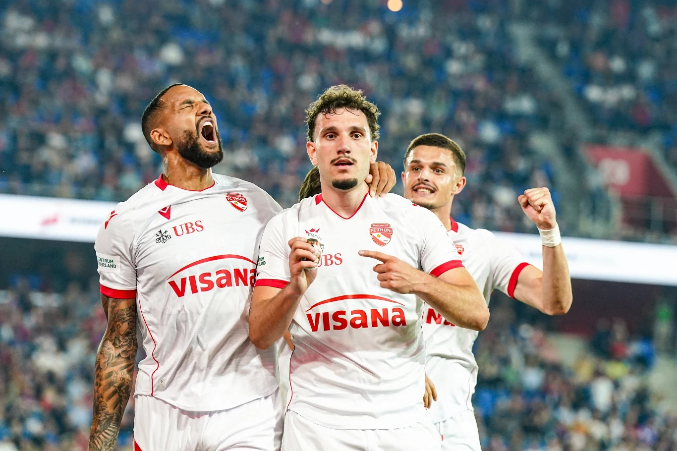 FC Thun mistrzem Szwajcarii Trzech piłkarzy FC Thun w białych strojach z logo VISANA i UBS świętuje gola na stadionie podczas meczu.