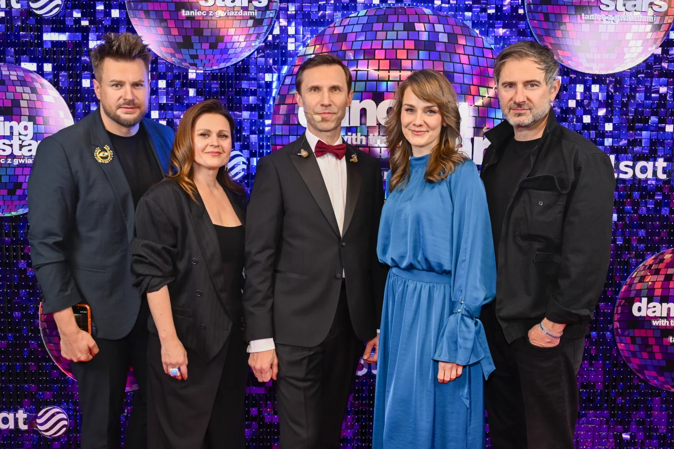 pięć osób pozuje razem na tle z motywem tanecznego show, wśród nich elegancko ubrany mężczyzna w garniturze z muchą, kobieta w niebieskiej sukience oraz pozostali w stonowanych kolorach, wszyscy patrzą w stronę obiektywu