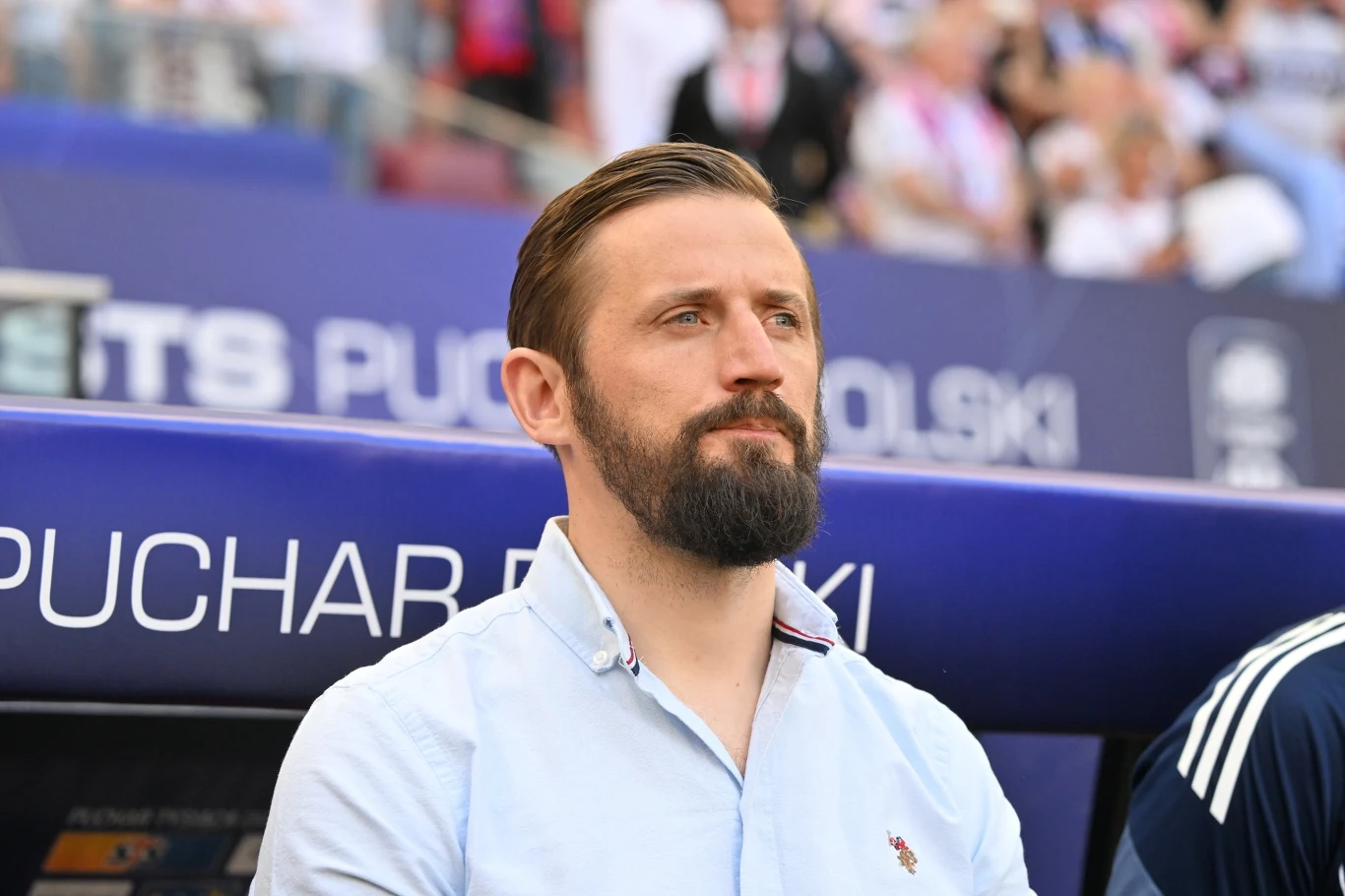 Łukasz Tomczyk mężczyzna z krótkimi, zaczesanymi do tyłu włosami i gęstą brodą, ubrany w jasną koszulę, siedzi na stadionowej ławce trenerskiej; w tle fragment baneru z napisem oraz rozmazany tłum widzów