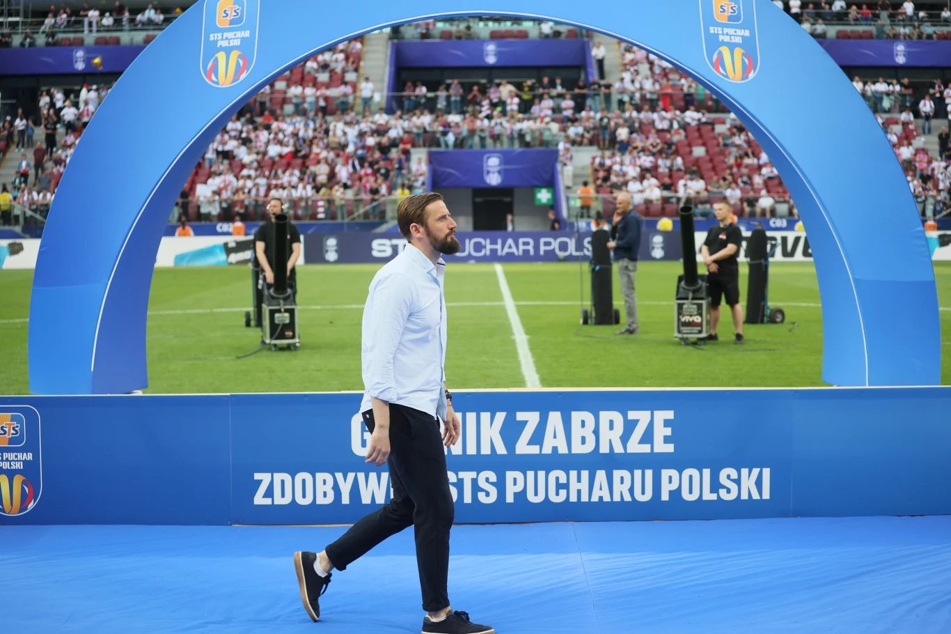 Łukasz Tomczyk mężczyzna ubrany w jasnoniebieską koszulę i ciemne spodnie przechodzi pod niebieskim łukiem z napisem informującym o zdobyciu Pucharu Polski przez Górnik Zabrze, w tle widać murawę stadionu, sprzęt telewizyjny oraz kibiców na trybunach