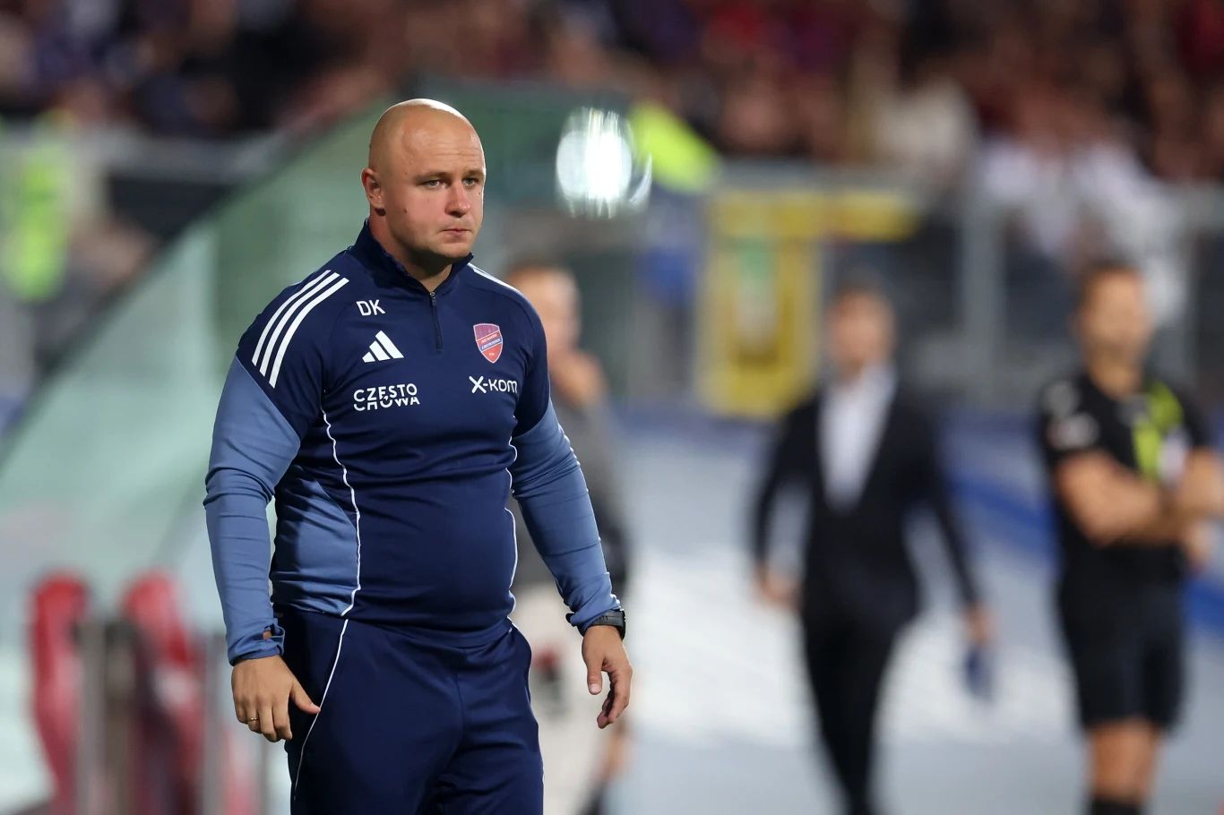 trener piłkarski w niebieskim dresie z logo klubowym stoi przy linii bocznej boiska, skupiony obserwuje zbliżającą się w powietrzu piłkę, w tle rozmyte sylwetki sędziego i innych osób na stadionie