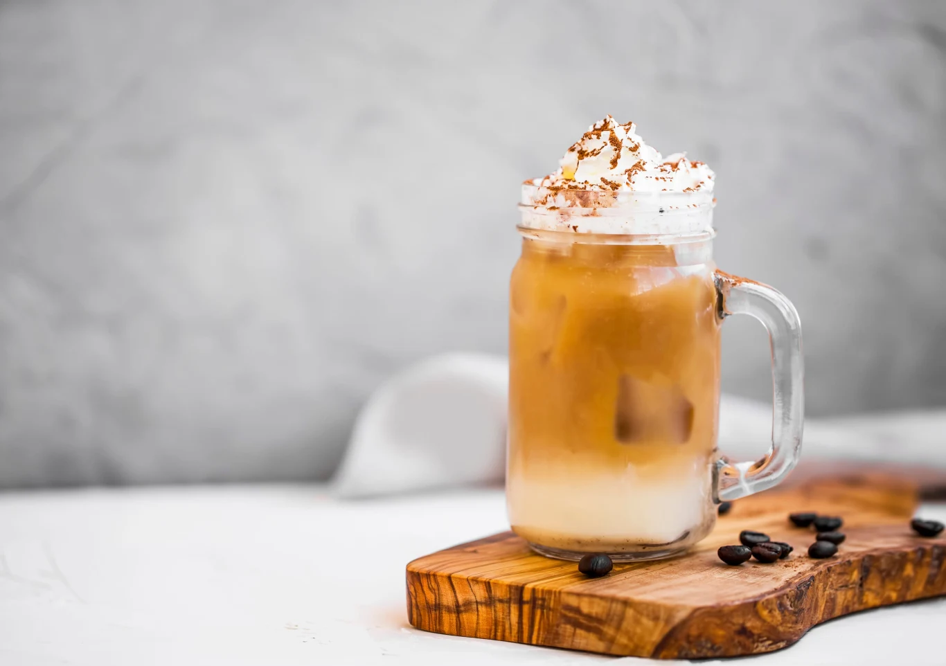 Bananową kawę możesz udekorować bitą śmietaną i wiórkami ciemnej czekolady Szklanka z mrożoną kawą latte z dużą ilością lodu i bitą śmietaną na wierzchu, posypaną kakao, ustawiona na drewnianej desce obok kilku ziaren kawy.