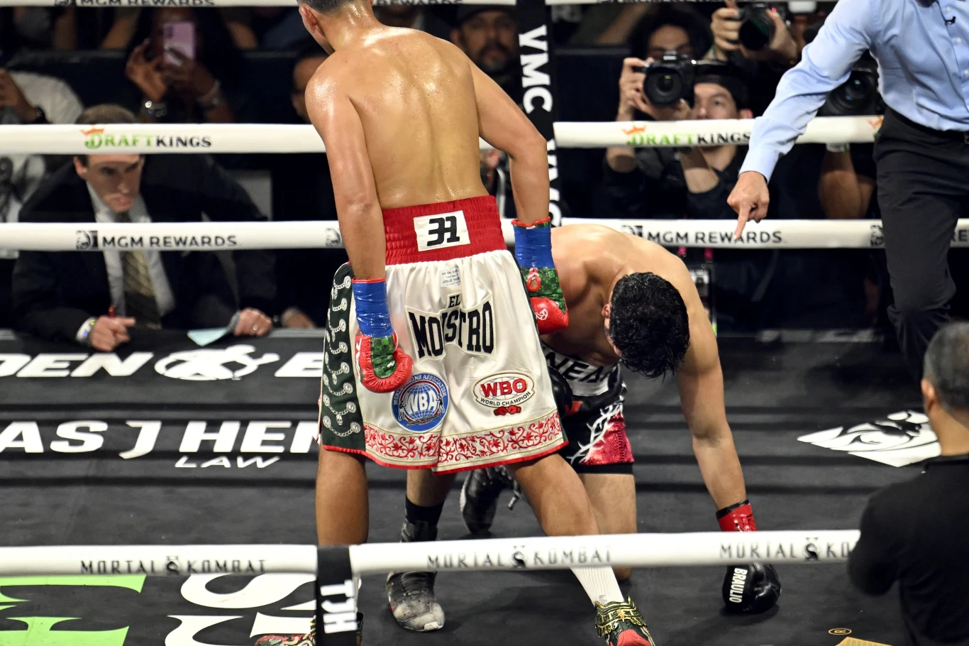 David Benavidez ma na deskach Gilberto Ramireza dwóch bokserów w ringu; jeden zawodnik stoi wyprostowany, drugi jest pochylony i opiera się o matę, sugerując nokaut lub silny cios, wokół nich fotografowie i sędzia obserwujący sytuację