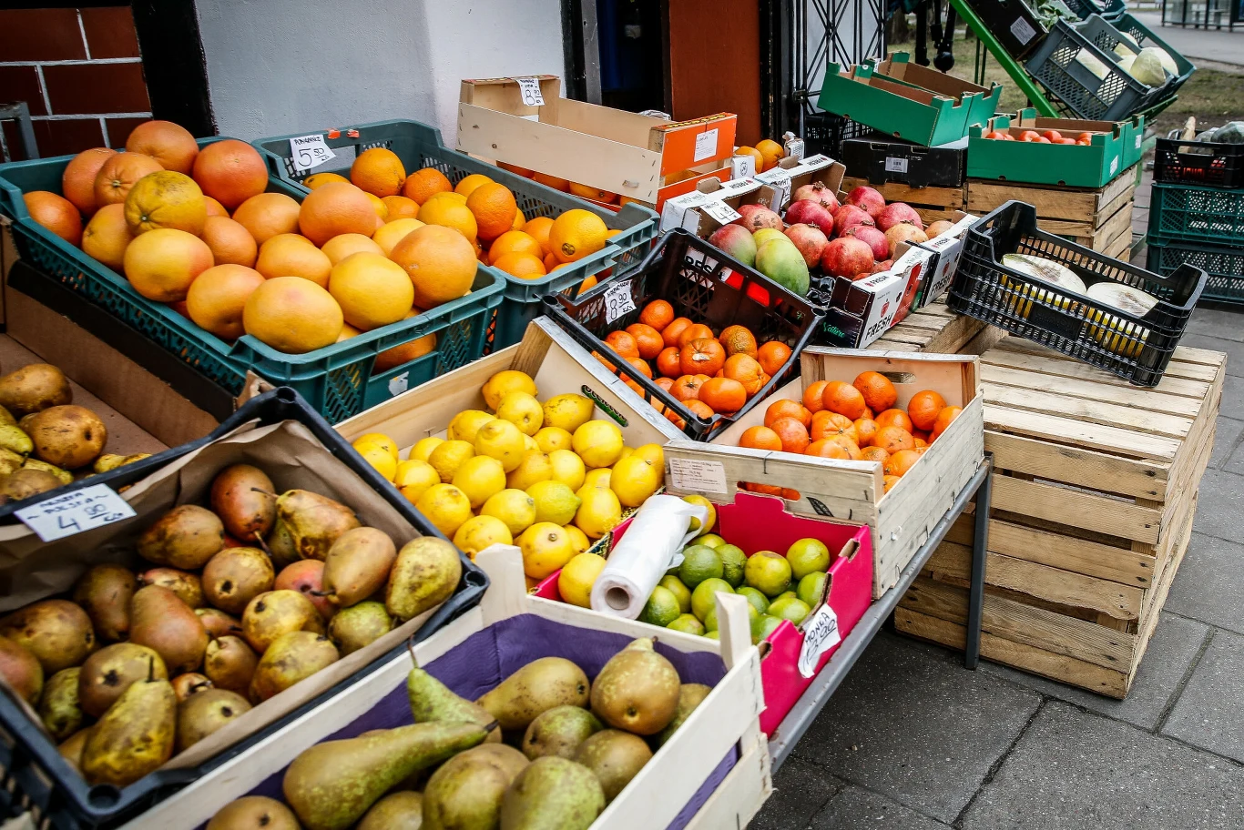 Różnorodne owoce ułożone w plastikowych i drewnianych skrzynkach, wśród których widać gruszki, cytryny, granaty, mandarynki, pomarańcze i pomelo, ustawione na stołach przed wejściem do sklepu warzywnego.