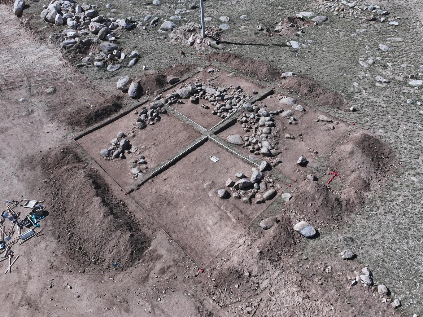 Kamienna ogrodzona konstrukcja obszar wykopalisk archeologicznych z wyraźnie wyznaczonymi kwadratowymi sektorami, wypełnionymi skupiskami kamieni oraz oddzielonymi pasami ziemi, wokół widoczne rozrzucone większe głazy na powierzchni